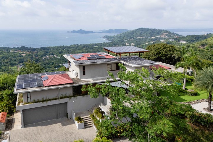 Modern hillside house with solar panels, overlooking ocean and green landscape.