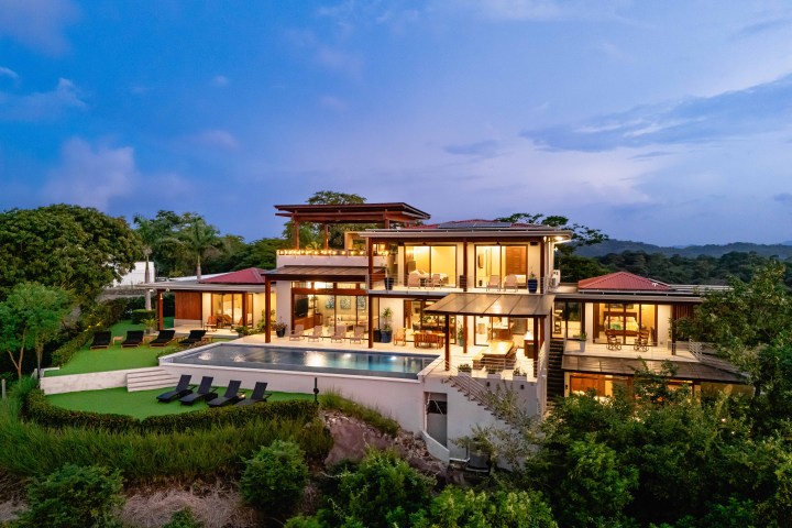 Modern hillside mansion with large windows, pool, and surrounding trees at dusk.