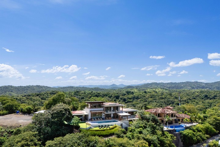 Aerial view of a large house with pool surrounded by lush greenery and hills under a clear blue sky.