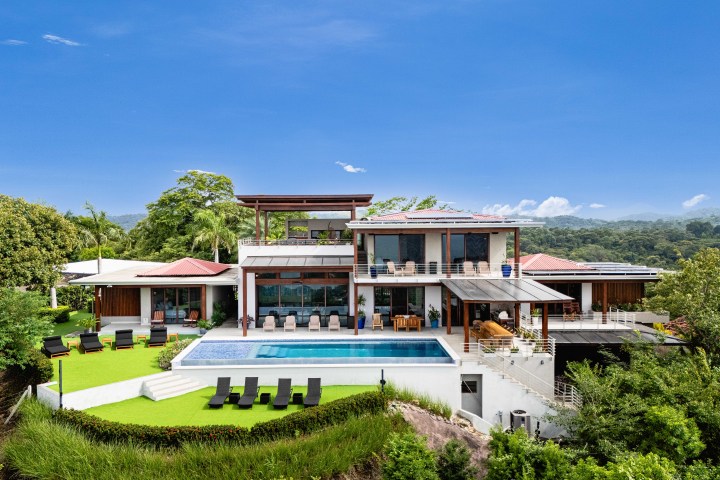 Modern house with large pool, surrounded by greenery, under a clear blue sky.