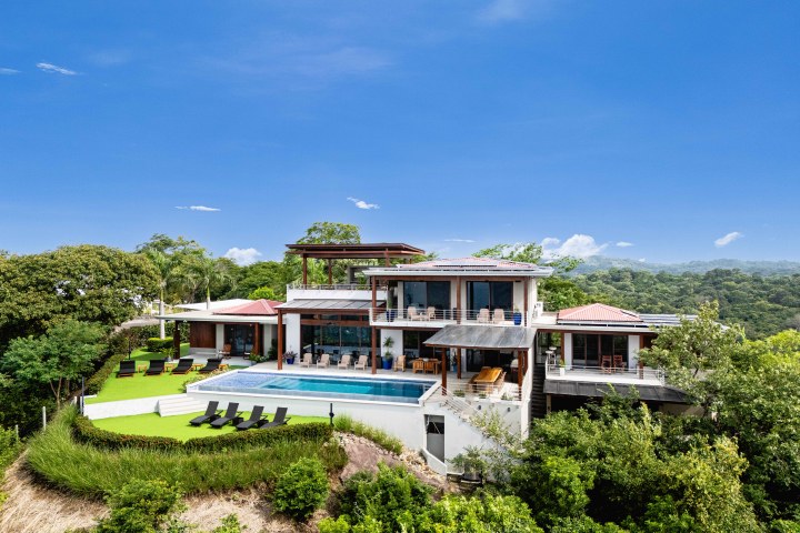 Modern hillside villa with pool and sun loungers, surrounded by trees under a clear blue sky.