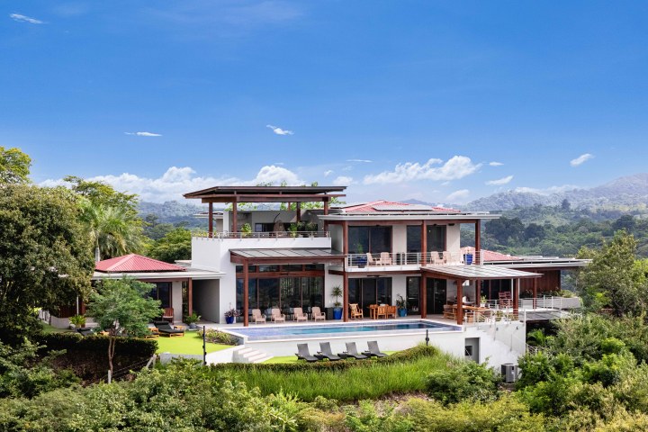 Modern hillside villa with a pool, surrounded by lush greenery and blue sky.