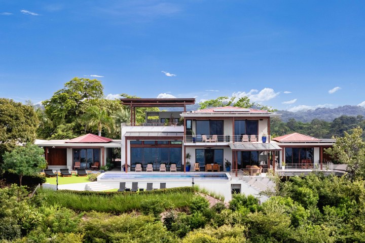 Modern villa with pool, surrounded by greenery and mountains, under a clear blue sky.
