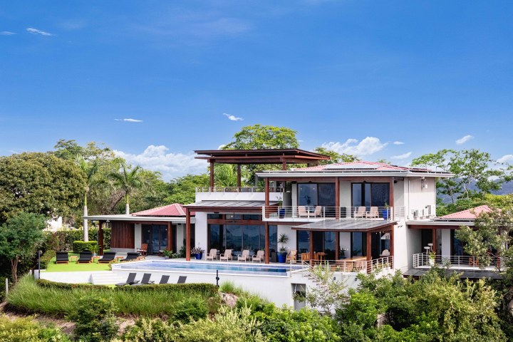 Modern two-story villa with a pool, surrounded by greenery and clear blue sky.