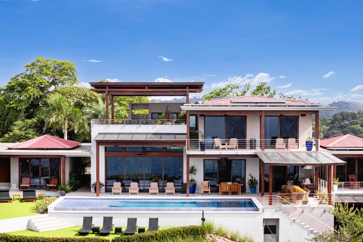 Modern two-story house with a pool and sun loungers, surrounded by trees.
