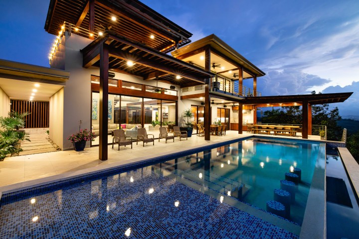 Modern house with large windows and a pool at dusk, surrounded by outdoor seating.