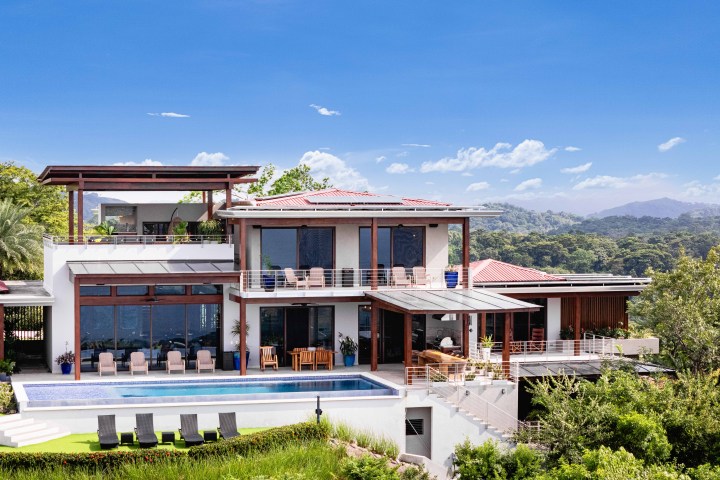Modern multi-level house with large windows, pool, and forested backdrop under a clear blue sky.