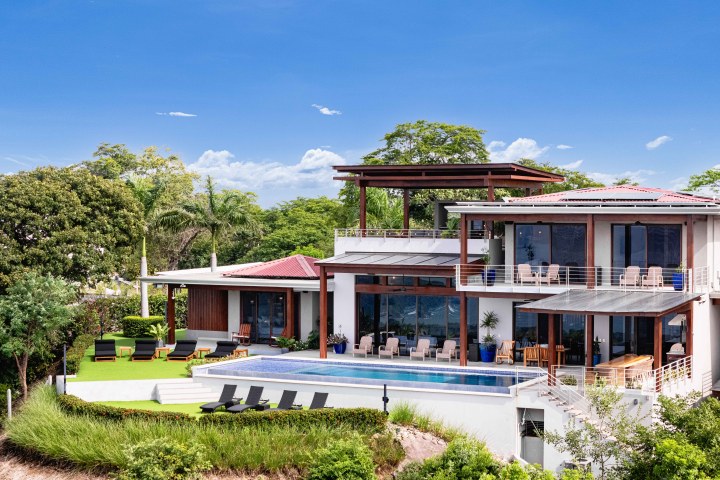 Modern two-story house with pool, surrounded by trees, under a clear blue sky.