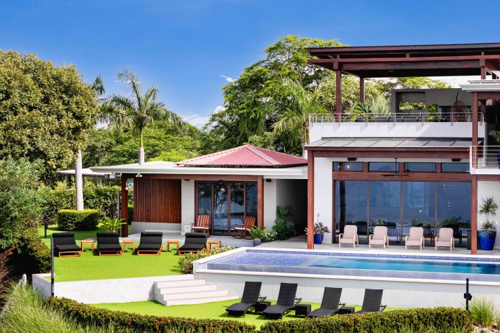 Modern villa with pool, green lawn, and palm trees in the background under a clear blue sky.