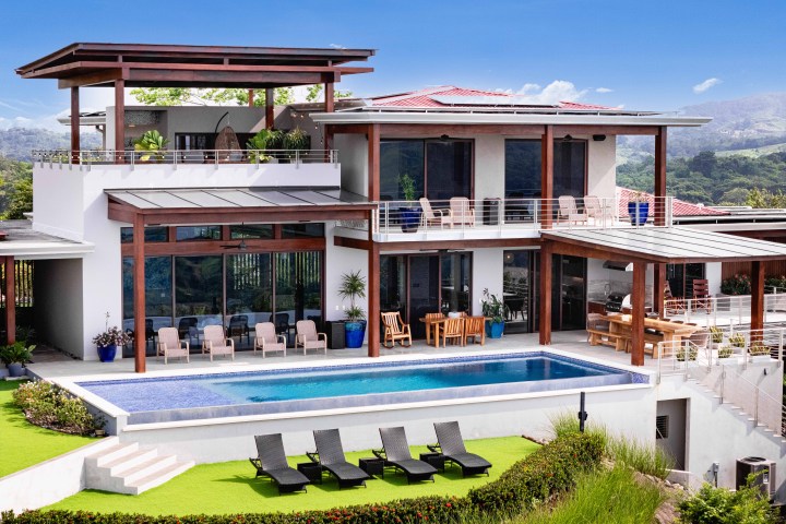 Modern two-story house with pool, patio furniture, and lush surroundings under a clear blue sky.