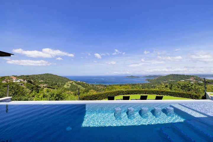 Infinity pool overlooking a lush hillside and ocean under a blue sky with scattered clouds.