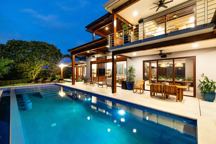 Modern house with a lit pool and patio at dusk, surrounded by trees.