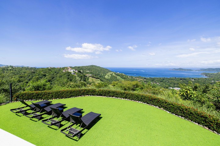 Lounge chairs on a grassy patio overlooking lush hills and a blue ocean under a clear sky.