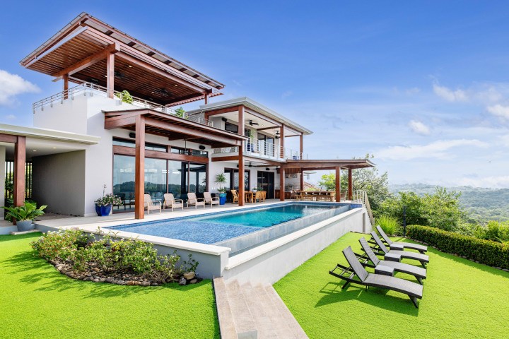 Modern hillside villa with a pool, deck chairs, and lush greenery under a clear blue sky.