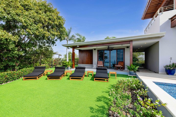 Modern villa with poolside lounge chairs on green lawn and lush garden.
