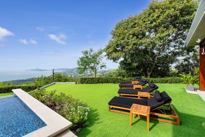 Poolside loungers on a grassy terrace with ocean and tree views under a blue sky.
