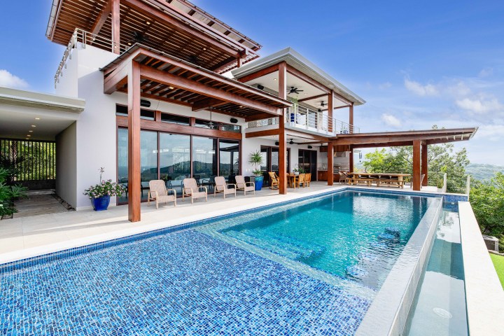 Modern house with pool and glass windows, surrounded by greenery under clear blue sky.