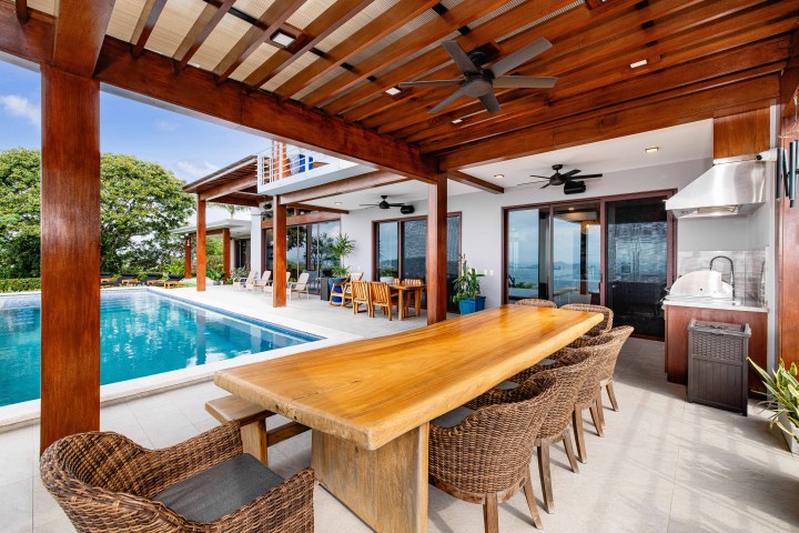 Outdoor dining area with wooden table, wicker chairs, and pool under shaded pergola.