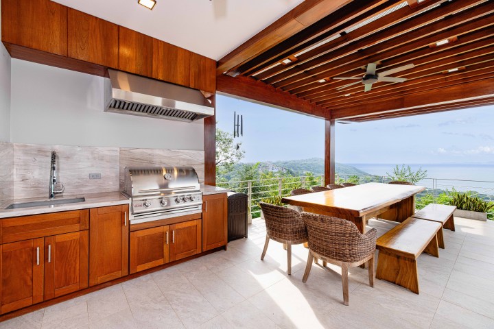 Outdoor kitchen with wooden dining area overlooking ocean and hills.