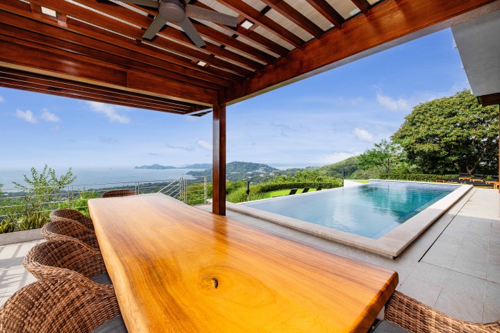 Wooden table under pergola with ocean view, adjacent to a rectangular pool and lush landscape.