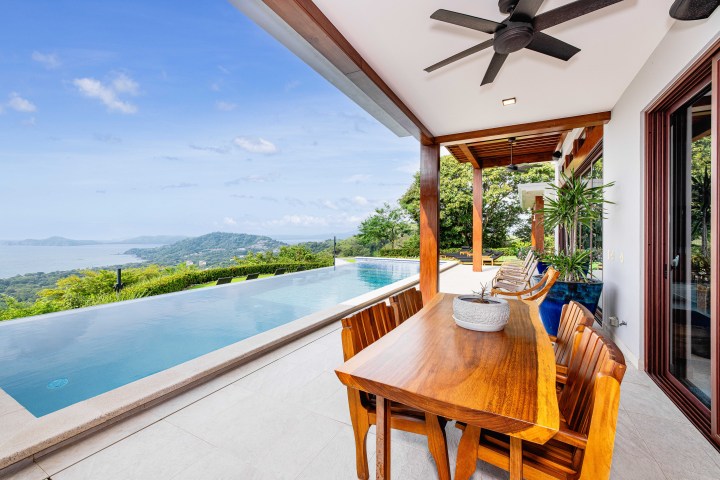 Patio with wooden table, chairs, ceiling fan, and infinity pool overlooking ocean and hills.