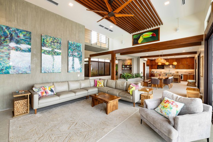 Modern living room with beige sofa, colorful pillows, artworks, and open kitchen view.