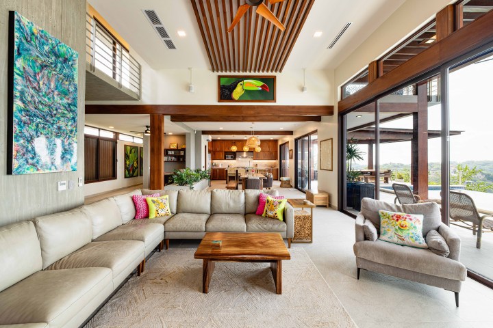 Spacious living room with beige couches, colorful pillows, artworks, large windows, and wooden ceiling accents.