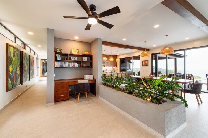 Modern open-concept living space with plants, ceiling fan, desk, bookshelves, and large windows with ocean view.