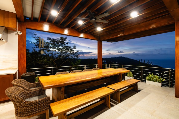 Outdoor dining area with wooden table, wicker chairs, and ocean view at sunset.