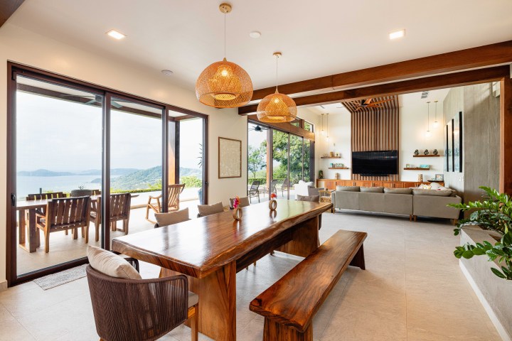 Open living area with wooden table, chairs, sofa, and large windows showing a scenic outdoor view.