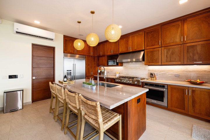 Modern kitchen with wooden cabinets, island with chairs, pendant lights, and stainless steel appliances.