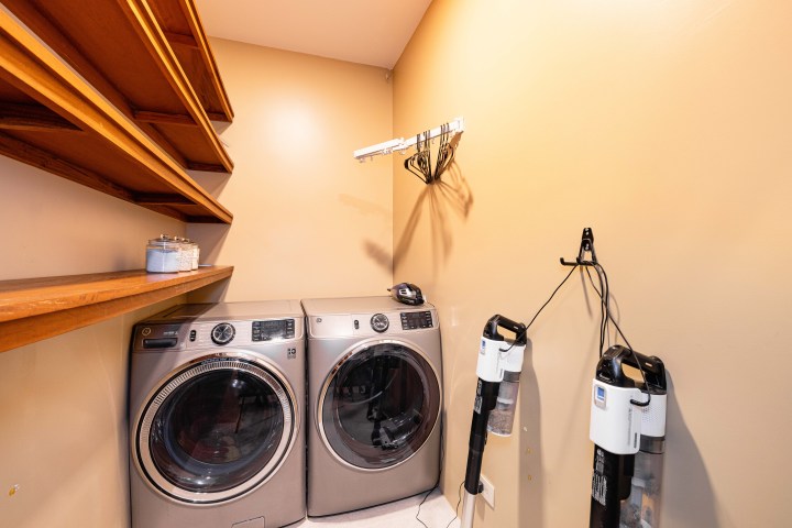 Laundry room with washer, dryer, wooden shelves, and wall-mounted vacuum cleaners.