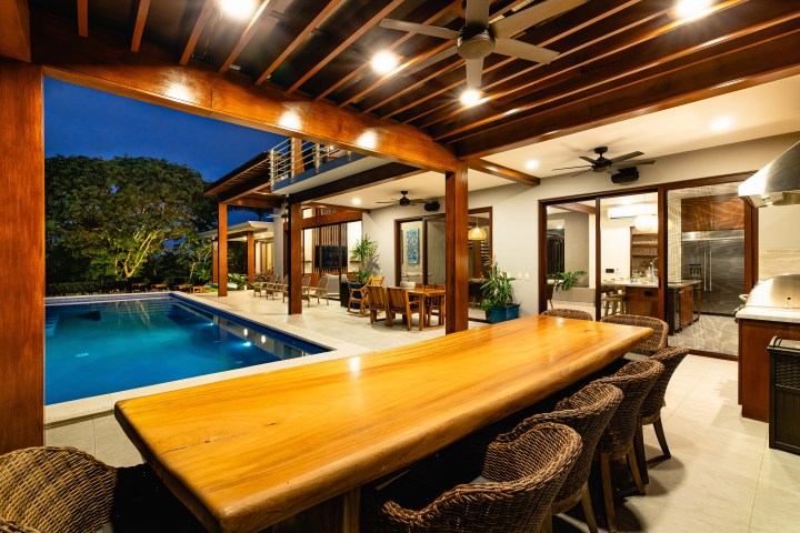 Outdoor dining area by a pool with a wooden table and wicker chairs, lit by overhead lights.
