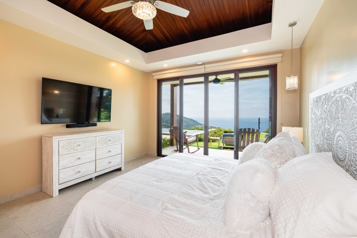 Bedroom with balcony ocean view, white bed, TV, dresser, and ceiling fan.