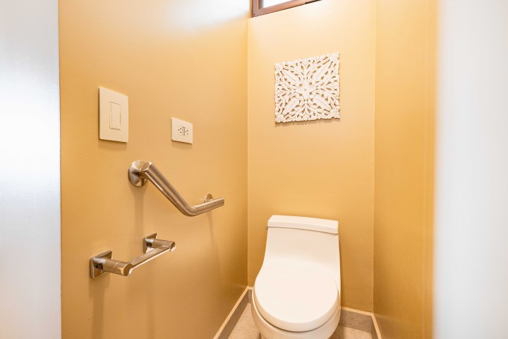 Small bathroom with toilet, beige walls, handrails, and decorative wall art.