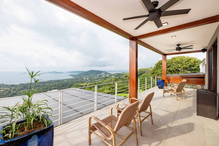Spacious terrace with chairs, ceiling fans, and ocean view surrounded by lush greenery and hills.