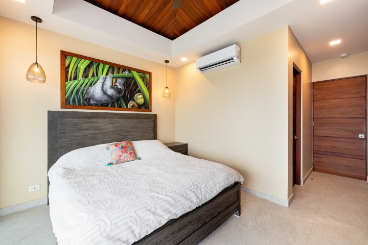 Bedroom with a wooden bed, white bedding, sloth painting, and ceiling fan.
