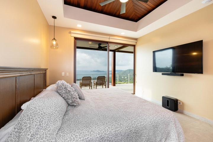 Bedroom with large window, bed, wall-mounted TV, and balcony overlooking sea and hills.