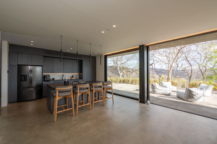 Modern kitchen with island, bar stools, and open view to patio with beanbag chairs and trees outside.