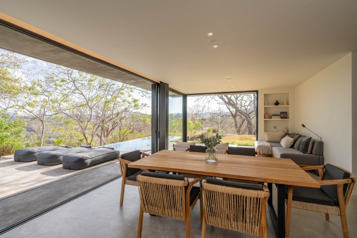 Modern living room with large windows, wooden table, gray sofa, and outdoor view of trees and patio.