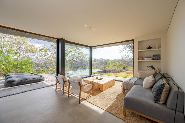 Modern living room with large windows, a sofa, chairs, and a view of greenery and a patio with cushioned seating.