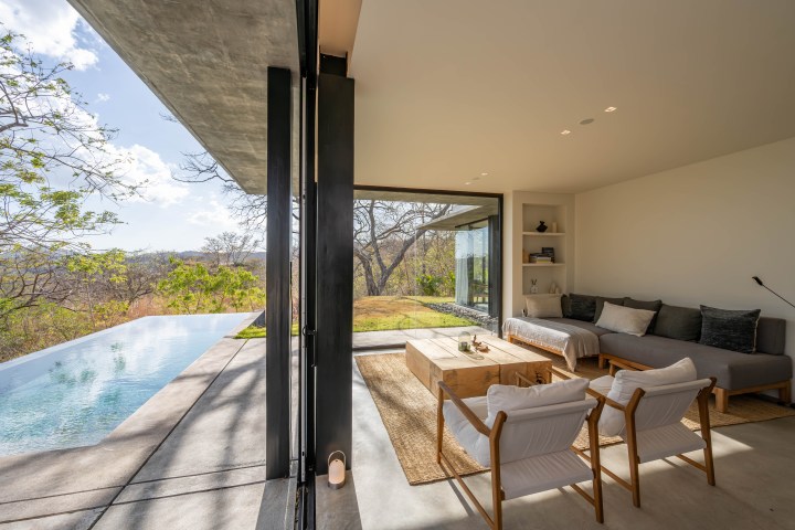 Modern living room with glass walls overlooking a patio and pool, featuring minimalist furniture.