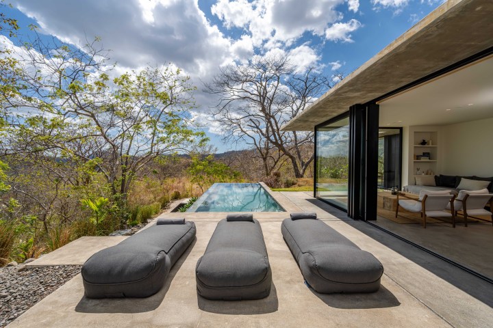 Modern patio with three loungers, pool, trees, and open living room under a blue sky with clouds.