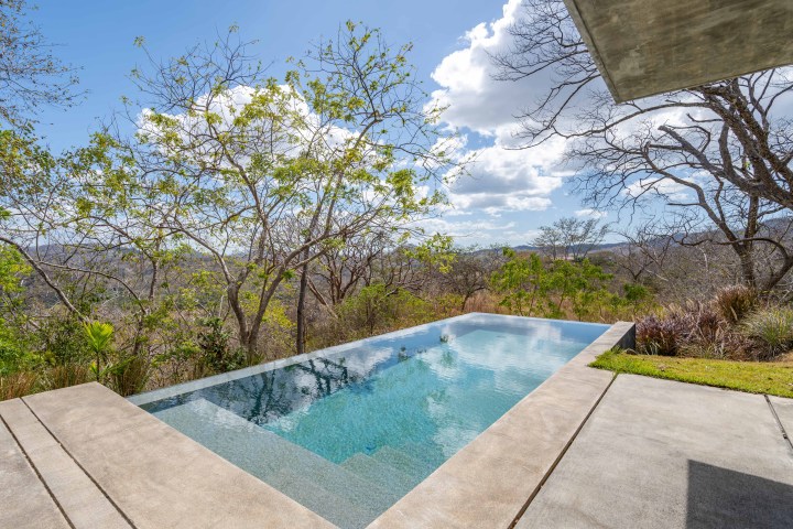 Infinity pool overlooking a landscape with trees and distant hills under a blue sky with clouds.