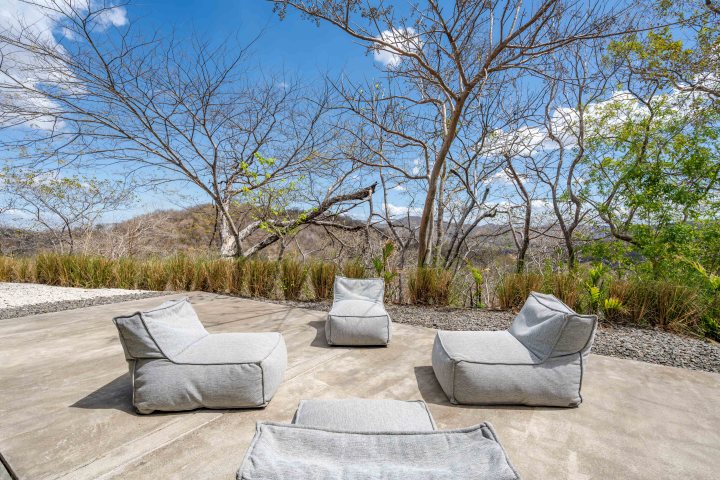 Outdoor seating area with gray bean bags on a patio overlooking bare trees and hills under a blue sky.