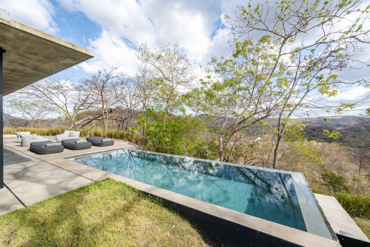 Infinity pool with lounge chairs overlooking a scenic hillside under a partly cloudy sky.
