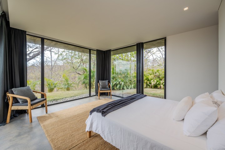 Modern bedroom with large windows, a bed, two chairs, and views of greenery.