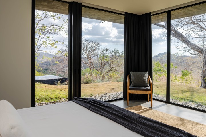 Bedroom with large windows showing a scenic outdoor view, a chair, and a bed in the foreground.