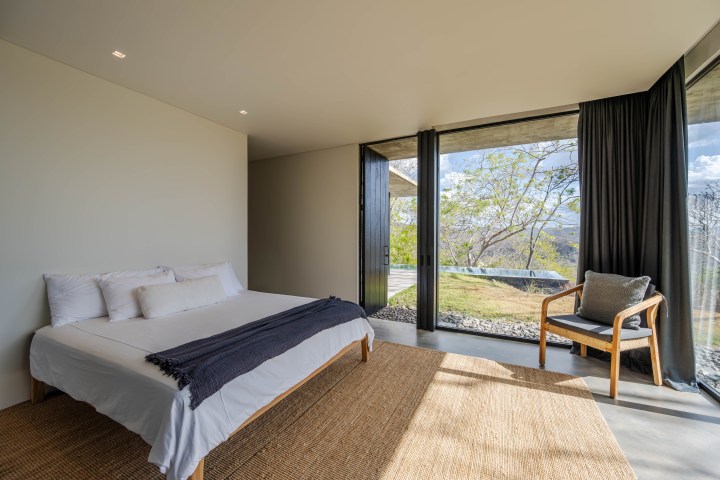 Minimalist bedroom with bed, chair, and large windows showing outdoor view.