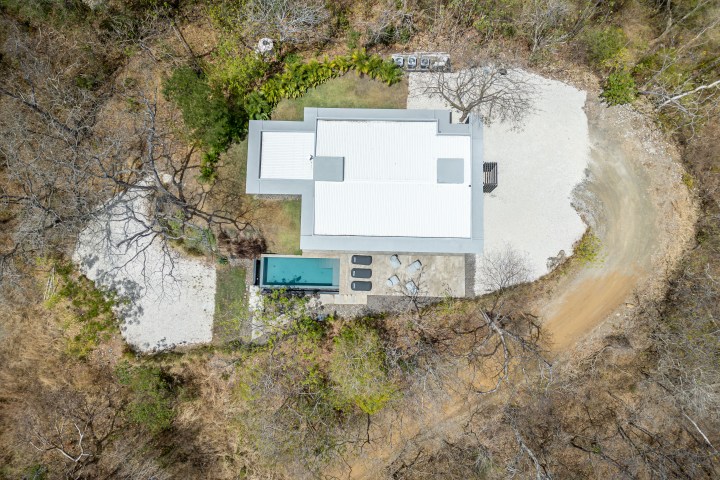 Aerial view of a modern house with a pool, surrounded by trees on a dirt road.